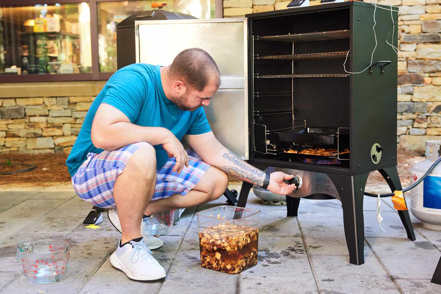 A person adjusts the dial of a gas smoker with a container of wood chips on the ground next to them.