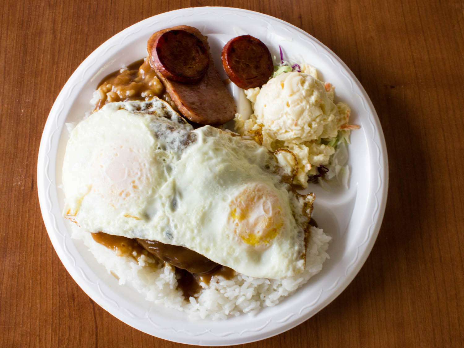 Overehad view of loco moco, served on a plate with a scoop of macaroni salad and griddled slices of Spam and Portuguese sausage.