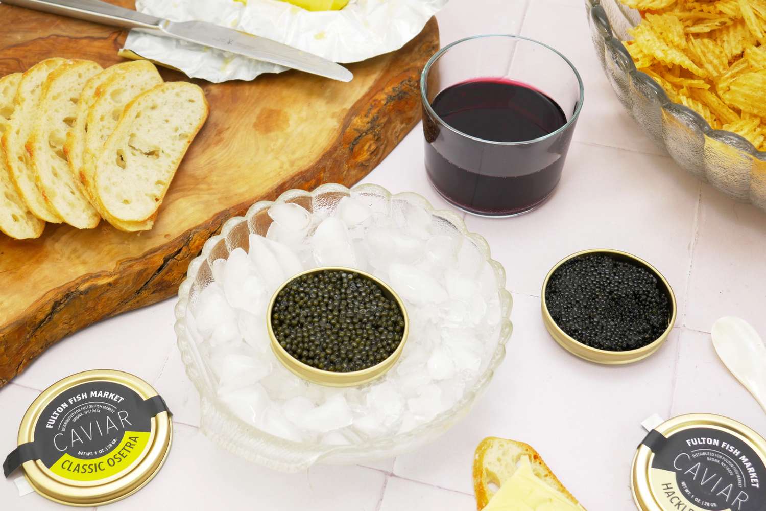A spread on a table featuring bread slices caviar tins with a bowl of ice chips and a glass of wine