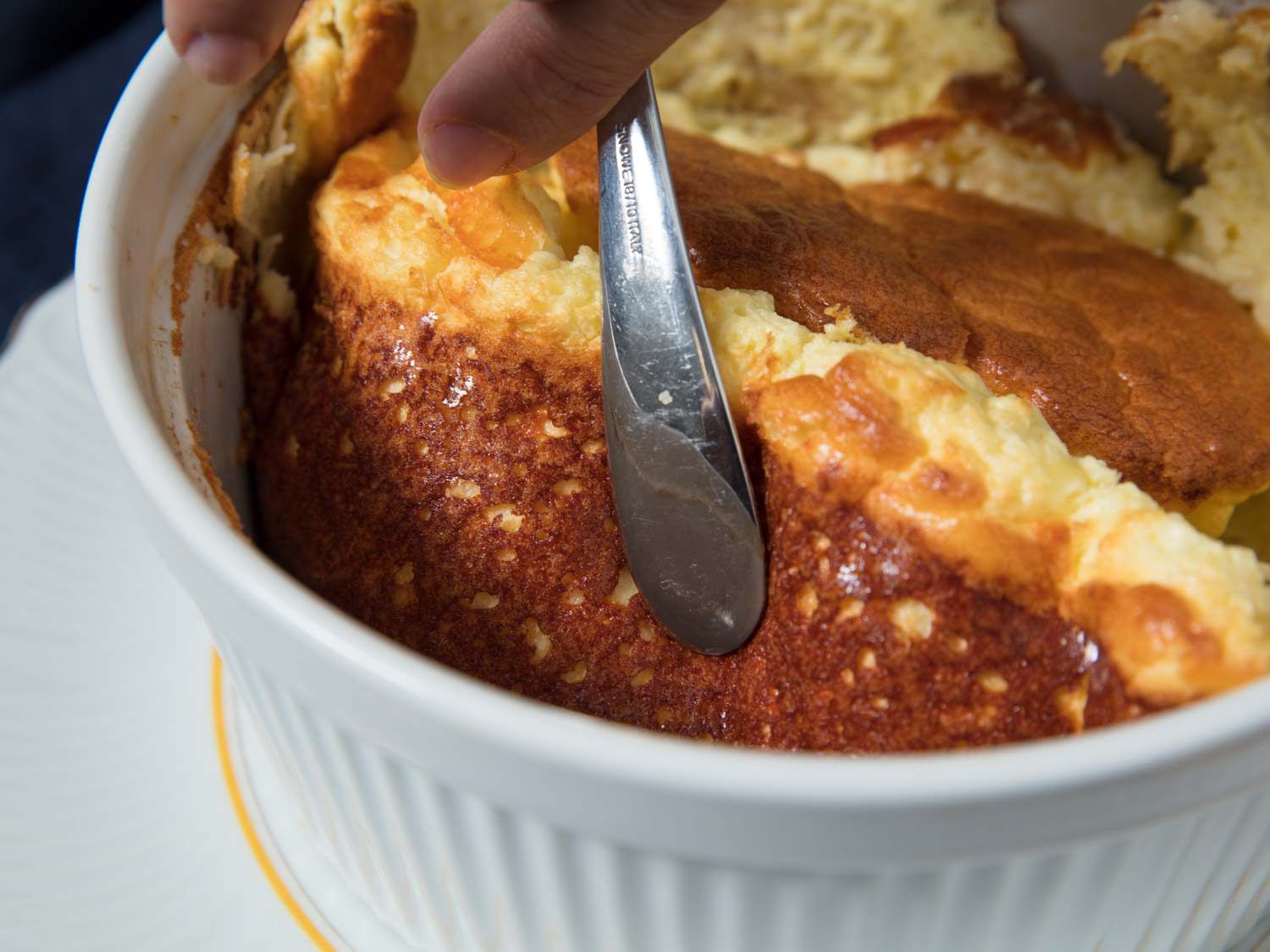 A spoon lifting the side of a souffle in a ramekin to show a beautiful, crispy crust after dusting the ramekin side with parmesan cheese.