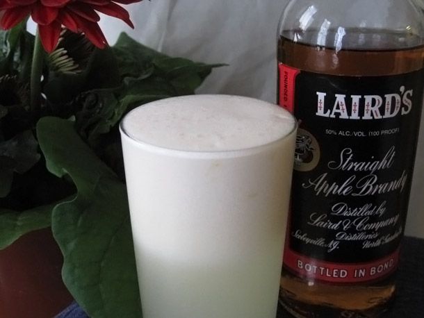 An apple blow fizz cocktail served in a pint glass. A bottle of apple brandy is nearby.