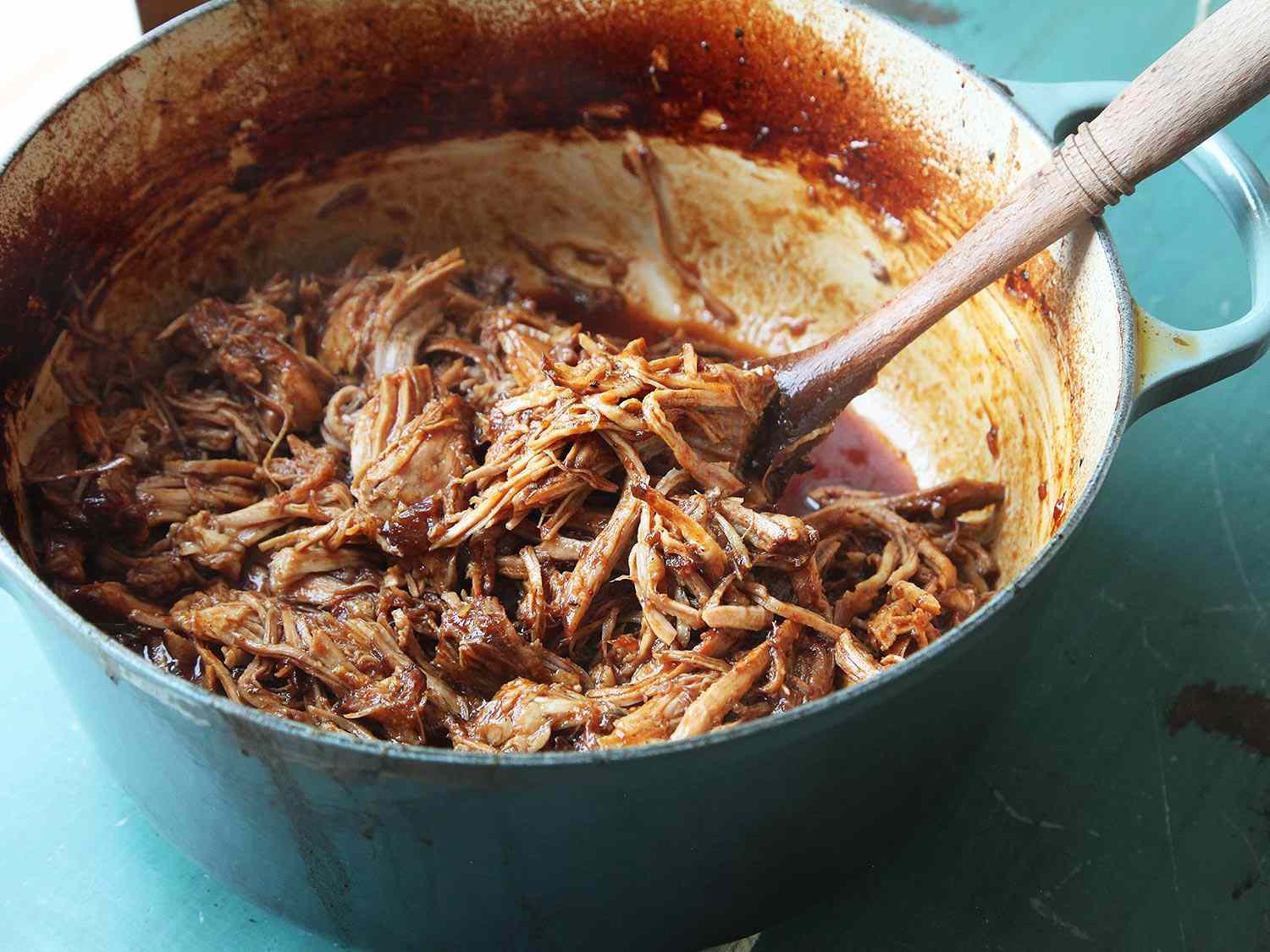 Pulled pork and a sauce being stirred together with a wooden spoon in a Dutch oven.