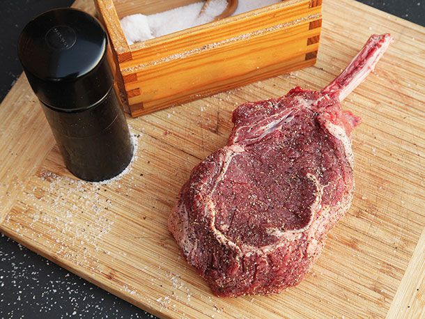 Raw bone-in bison ribeye steak seasoned with salt and pepper on a wooden board.