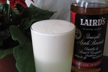 An apple blow fizz cocktail served in a pint glass. A bottle of apple brandy is nearby.