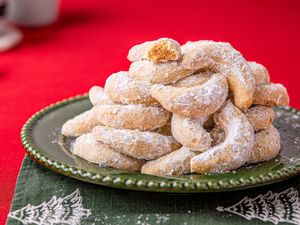 A stack of crescentshaped cookies dusted with powdered sugar arranged on a green plate placed over a festive napkin