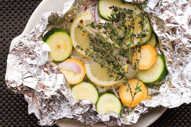 Overhead shot of green and yellow zucchini slices, sliced red onion, and thyme sprigs layered over cod in an open foil packet 