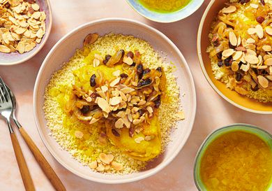 2 plates of Chicken couscous T'faya on pink surface surrounded by bowls of broth, toasted almonds, and forks