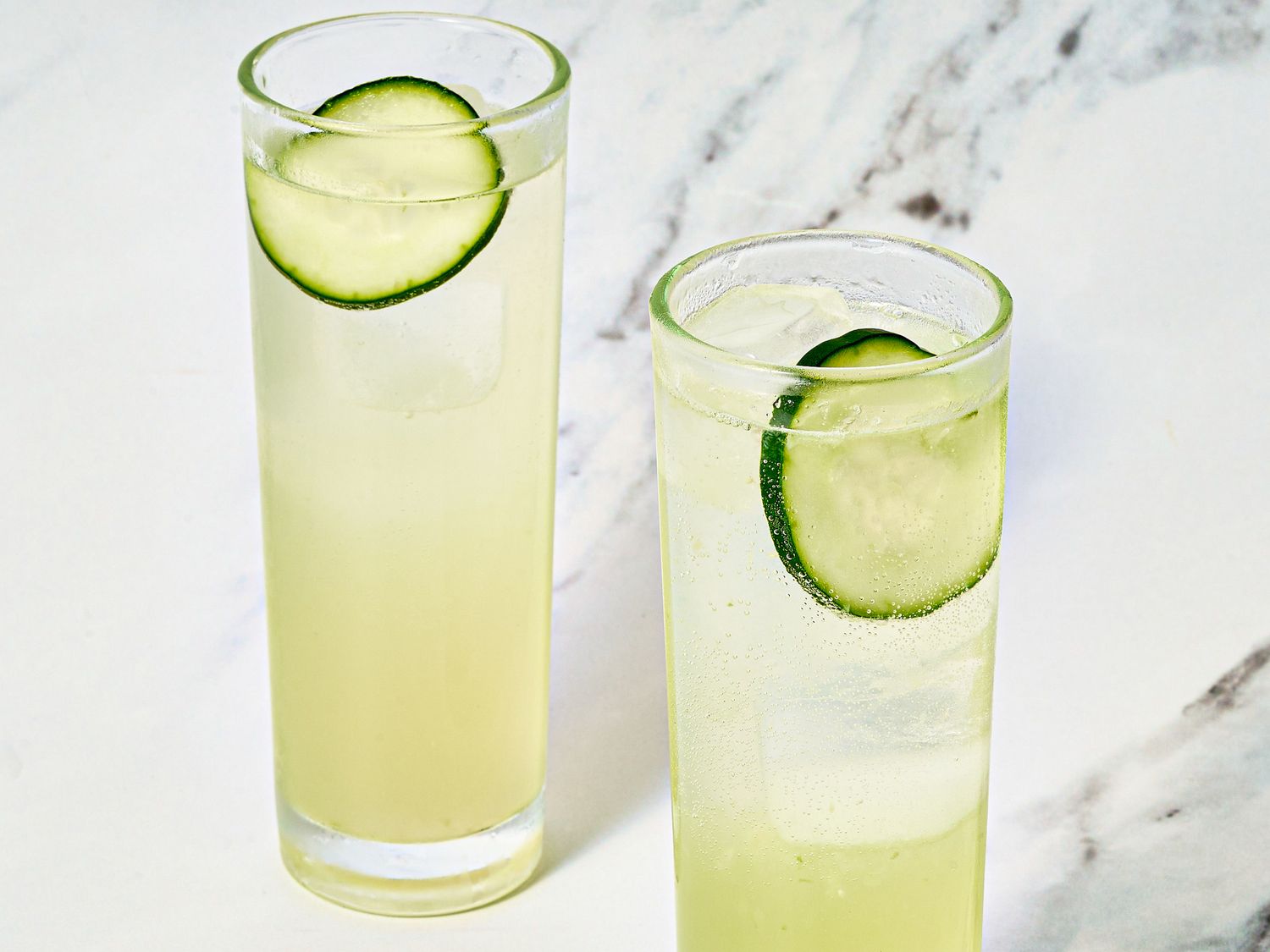 Two glasses with cucumber garnished beverages on a marble surface