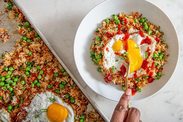 A serving of fried rice with peas and a fried egg on top, being scooped with a spoon from a white bowl