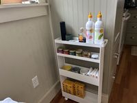 A white rolling kitchen storage rack with olive oil, spices, mugs, napkins, and boxes of pasta on its shelves