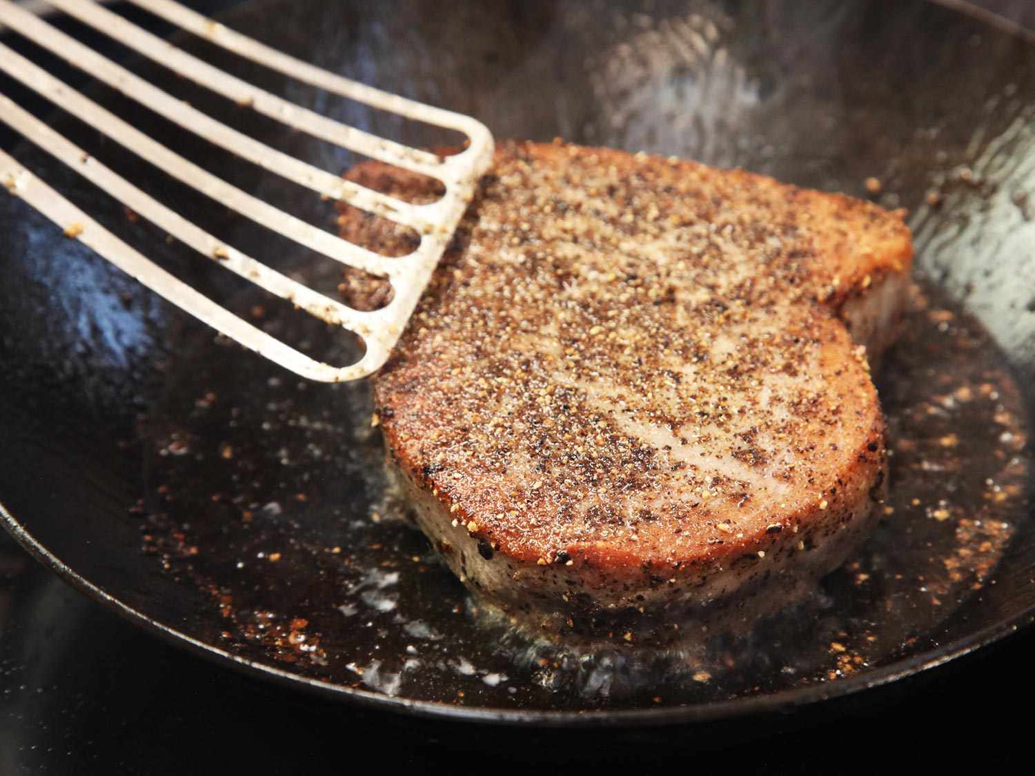 Tuna steak has just been turned with a fish spatula to sear on its second side. The side facing up has an attractive browned surface.