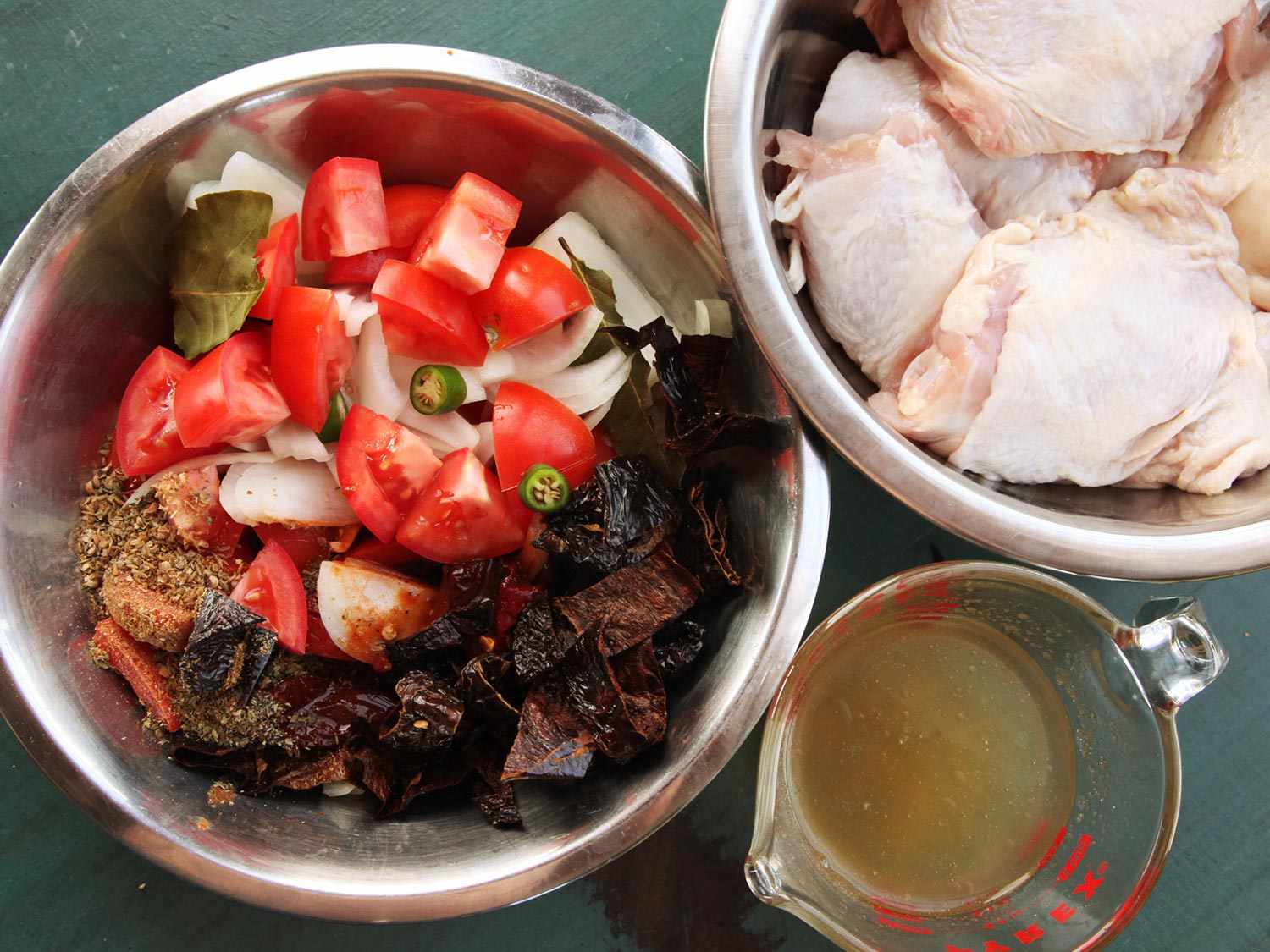 Overhead view of enchilada ingredients: a measuring cup of stock, a bowl of raw chicken pieces, and a bowl of vegetables, chiles, and seasonings.