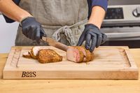 Person slicing roasted meat on a John Boos Reversible Maple Wood Cutting Board with Juice Groove wearing gloves and an apron kitchen countertop in background