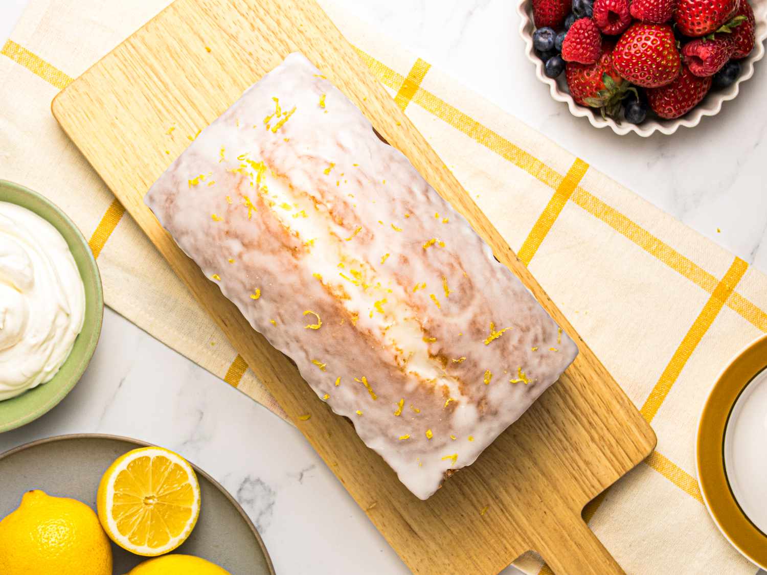 Lemon pound cake with icing and lemon zest on a wooden board, surrounded by lemons, whipped cream, and mixed berries in bowls.