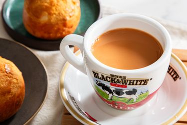 Cup of milk tea in a logoed mug on a plate with baked goods in the background