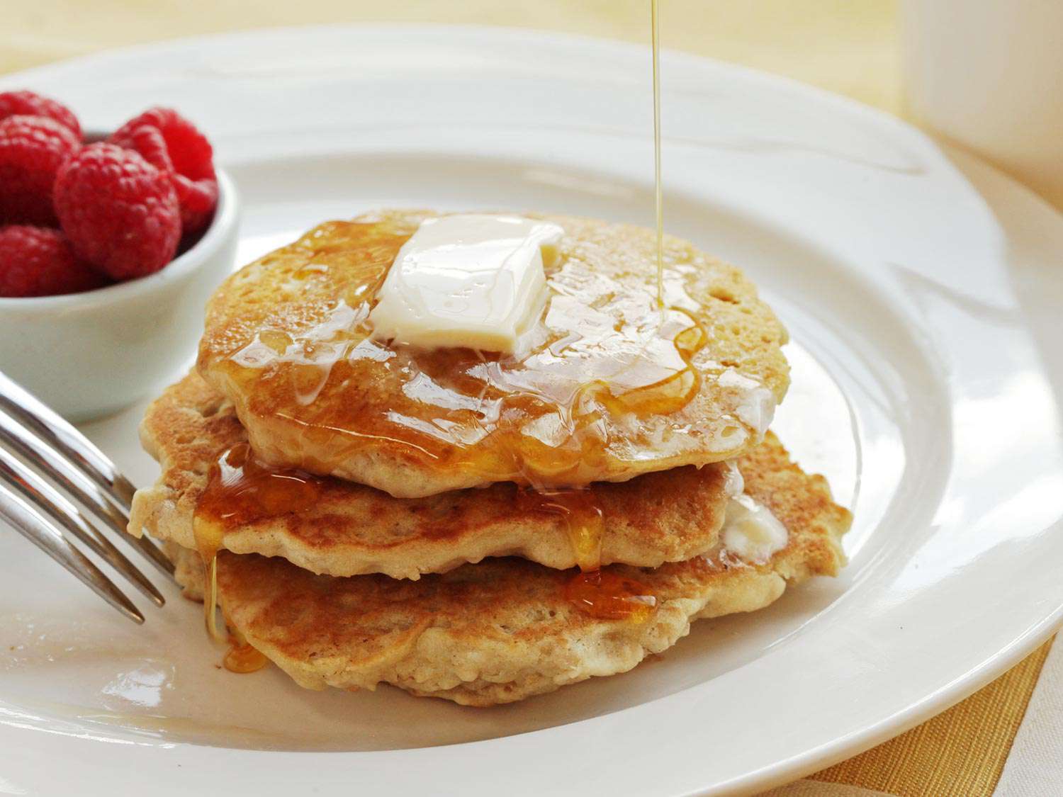 A stack of vegan oatmeal pancakes with syrup and butter. A small dish of raspberries is next to the pancakes. 