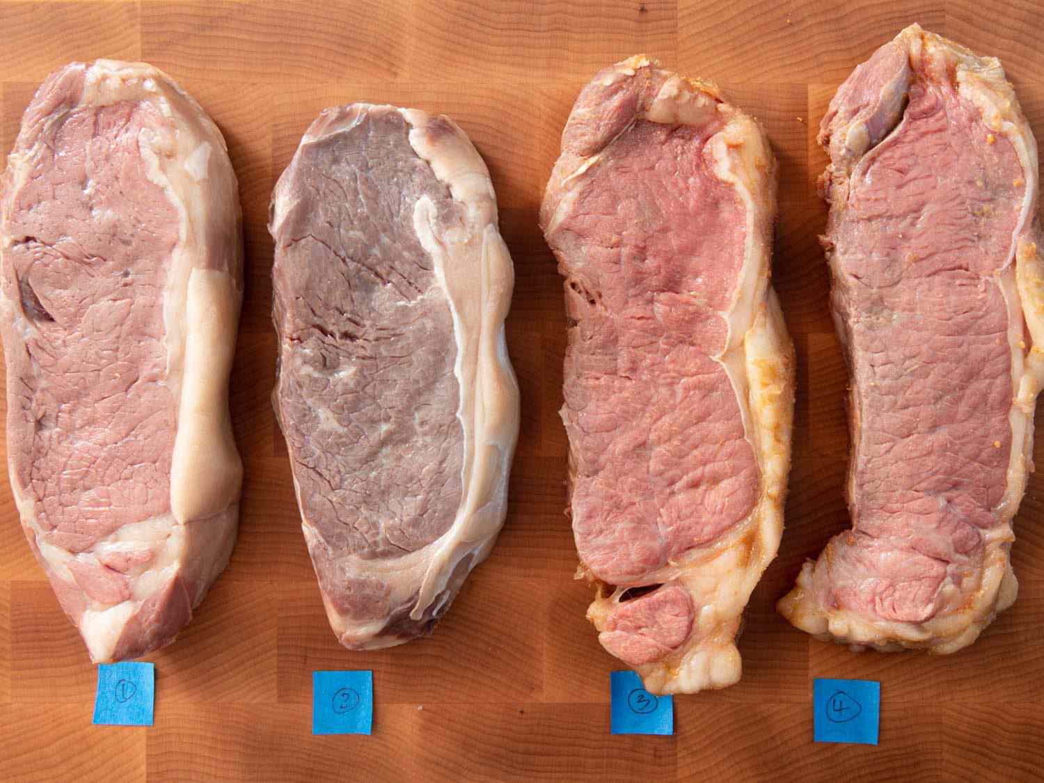 Overhead view of four strip steaks cooked sous vide, identified by numbered tape on a cutting board. The second steak from the left is considerably grayer than the other three.
