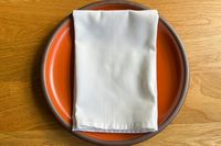 A white napkin neatly folded on an orange plate set on a wooden table