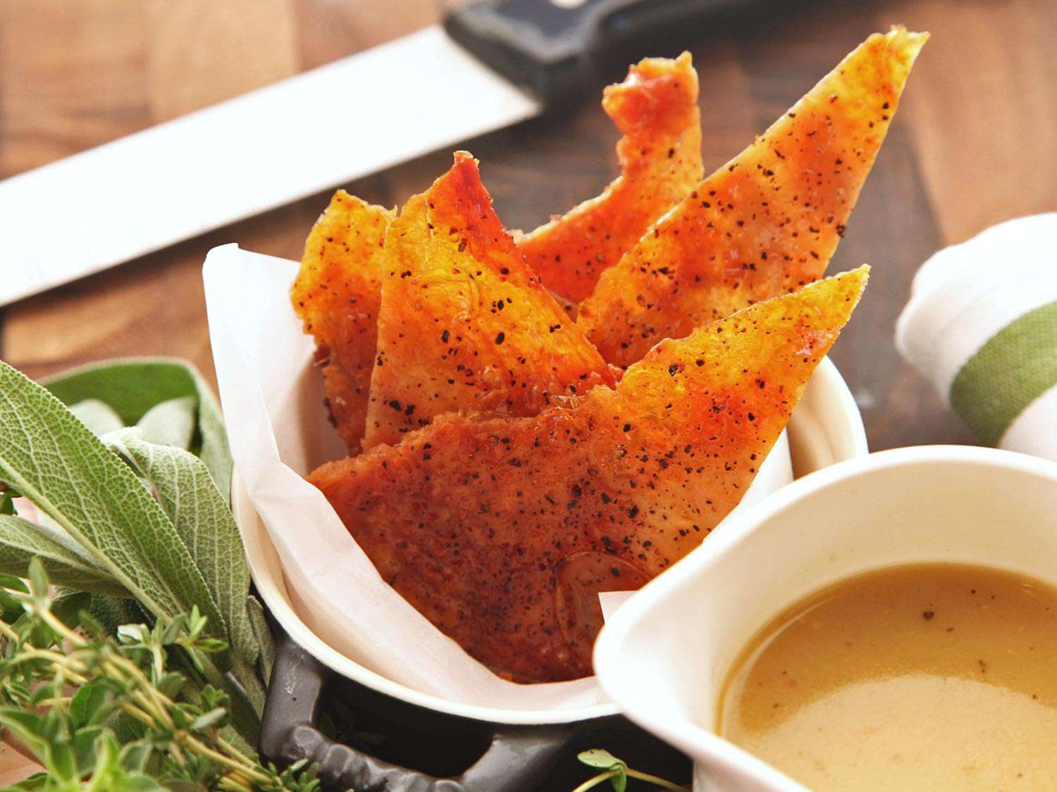 Container of crisp turkey skin shards next to herb garnish and gravy boat.