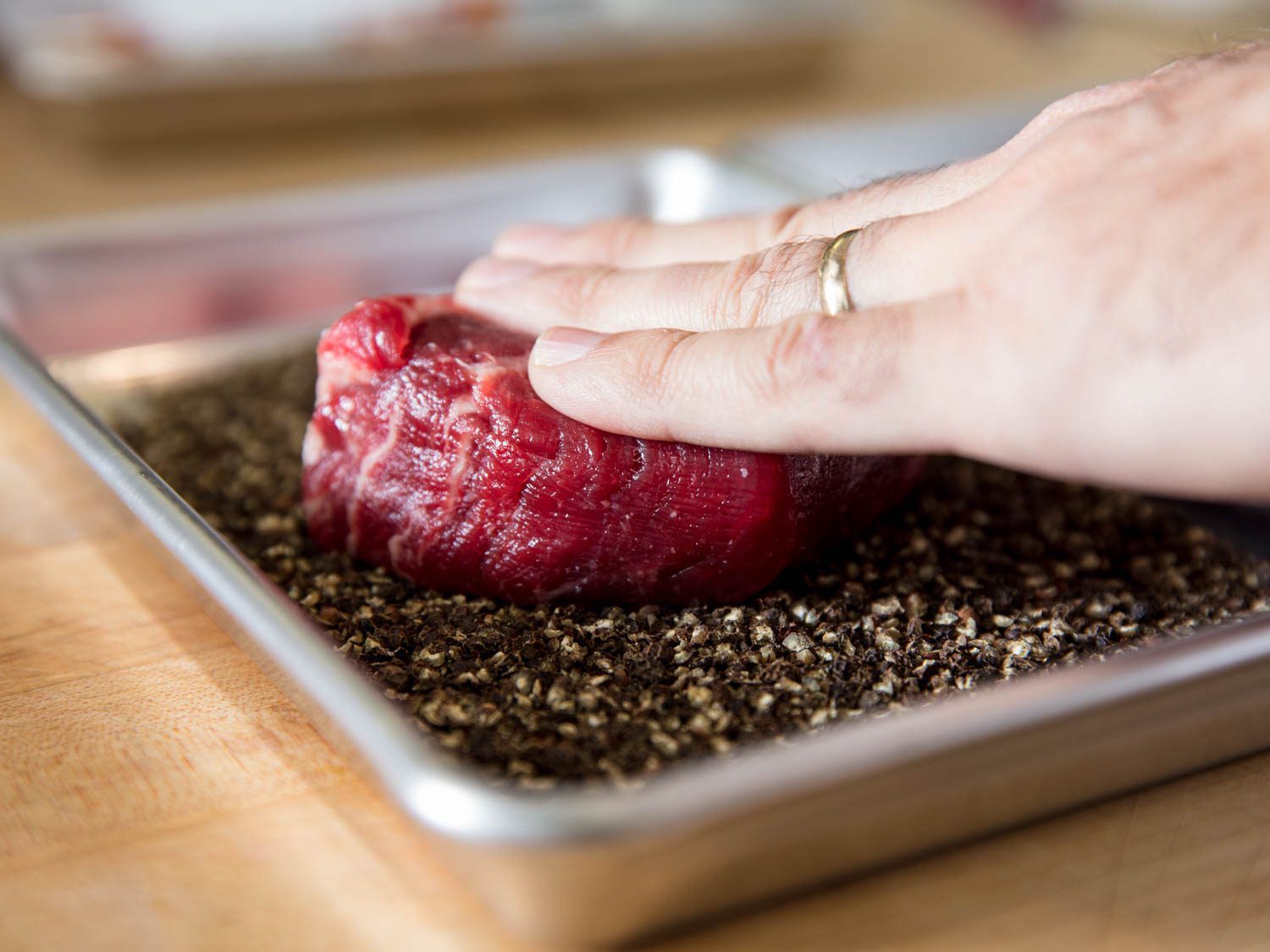 Steaks are placed into a rimmed baking sheet containing the cracked peppercorns and pressed so that they adhere.
