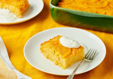A slice of orange-flavored cake served on a white plate with a dollop of cream and a fork, with the rest of the cake in a green dish in the background