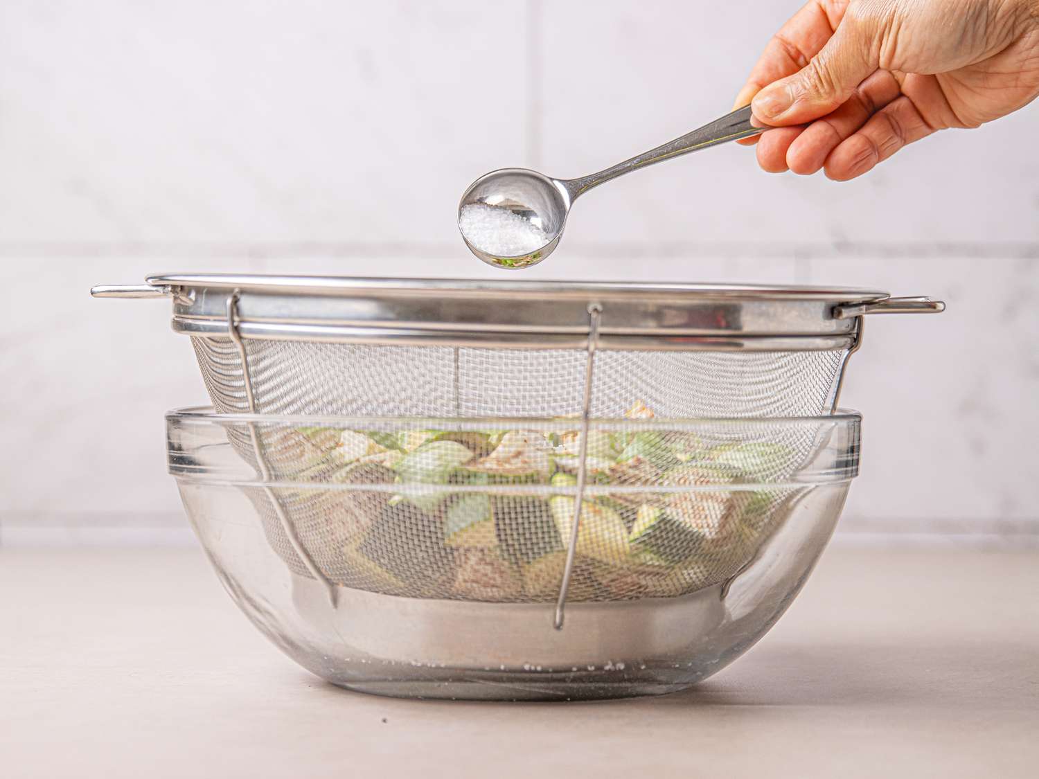 A strainer placed over a glass bowl containing chopped vegetables a person adding salt with a spoon