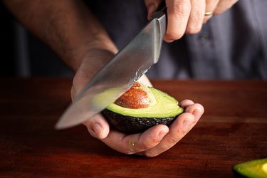 A person removing the pit from half of an avocado with a knife closeup on hands and the avocado