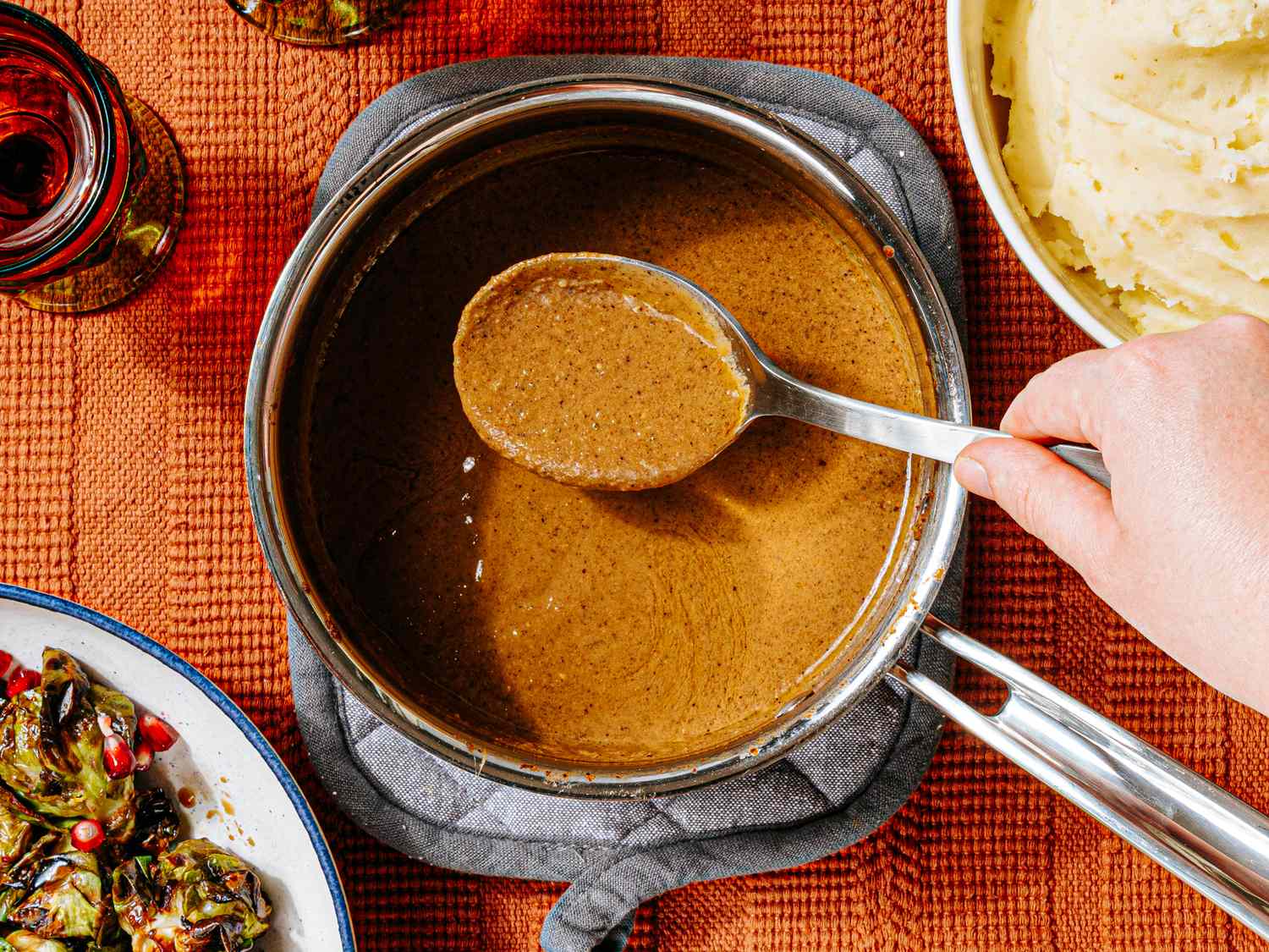 A person scooping vegan gravy from a pot with a ladle with mashed potatoes and vegetables visible nearby