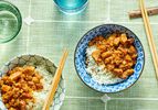 2 bowls of minced pork on white rice in a blue bowl and a white bowl, with chop sticks and glasses of water on a green tablecloth