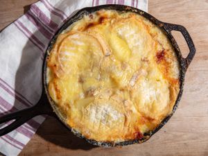 A cast iron skillet full to the brim with baked potato tartiflette, topped with melted and browned soft-rind cheese.