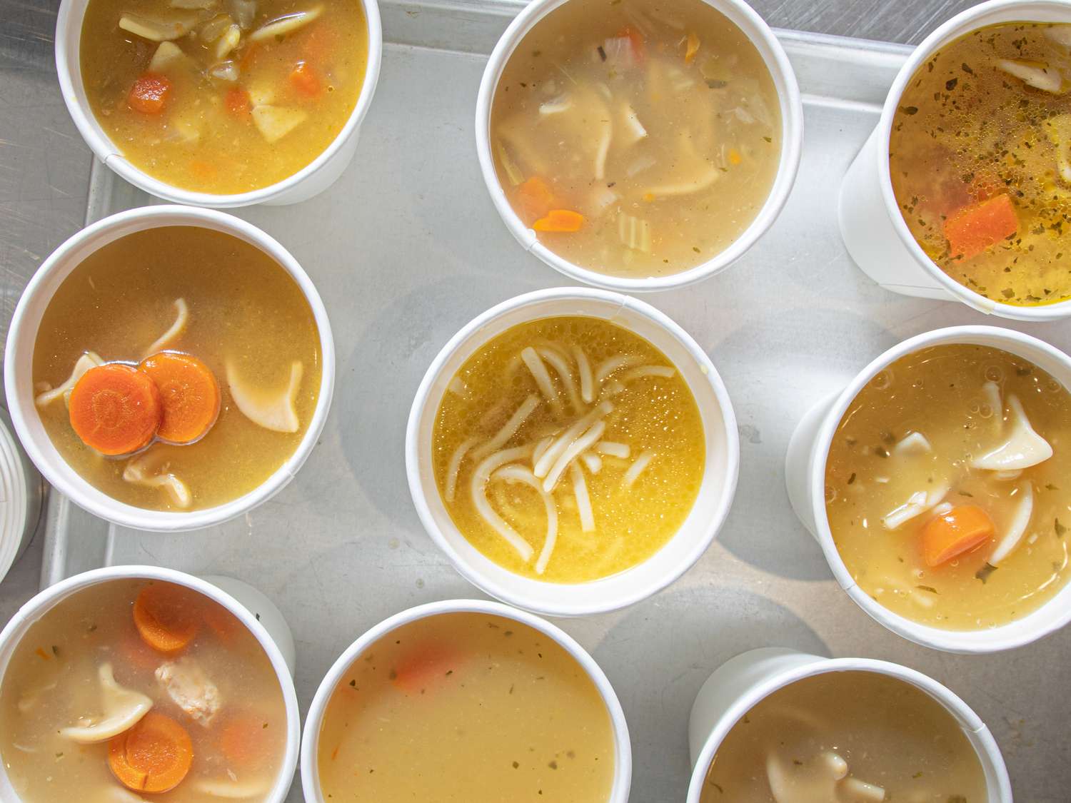 Various cups of chicken noodle soup on a tray showing variety in ingredients and presentation