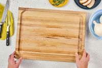 Hands holding a John Boos Reversible Maple Wood Cutting Board with Juice Groove on a kitchen counter surrounded by knives a towel onions rosemary and sliced meat