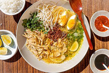 Bowl of Soto Ayam with wooden spoon, surrounded by limes, sambal, soy sauce and rice on a bamboo matt