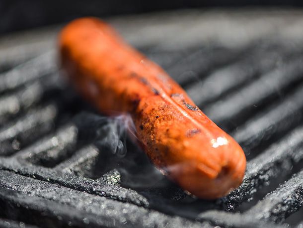 Close-up of a skinless hot dog getting browned on a smoking grill grate.