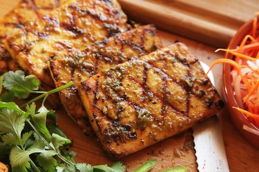 Four slabs of grilled tofu with appetizing grill marks, next to fresh cilantro and shaved carrots.