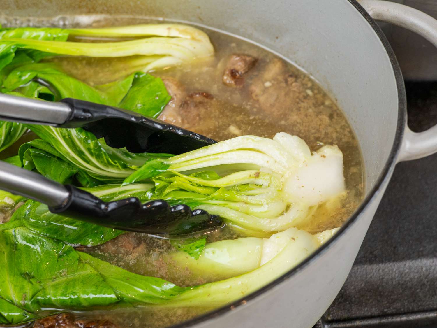 Tongs lifting bok choy out of oxtail broth in a Dutch oven
