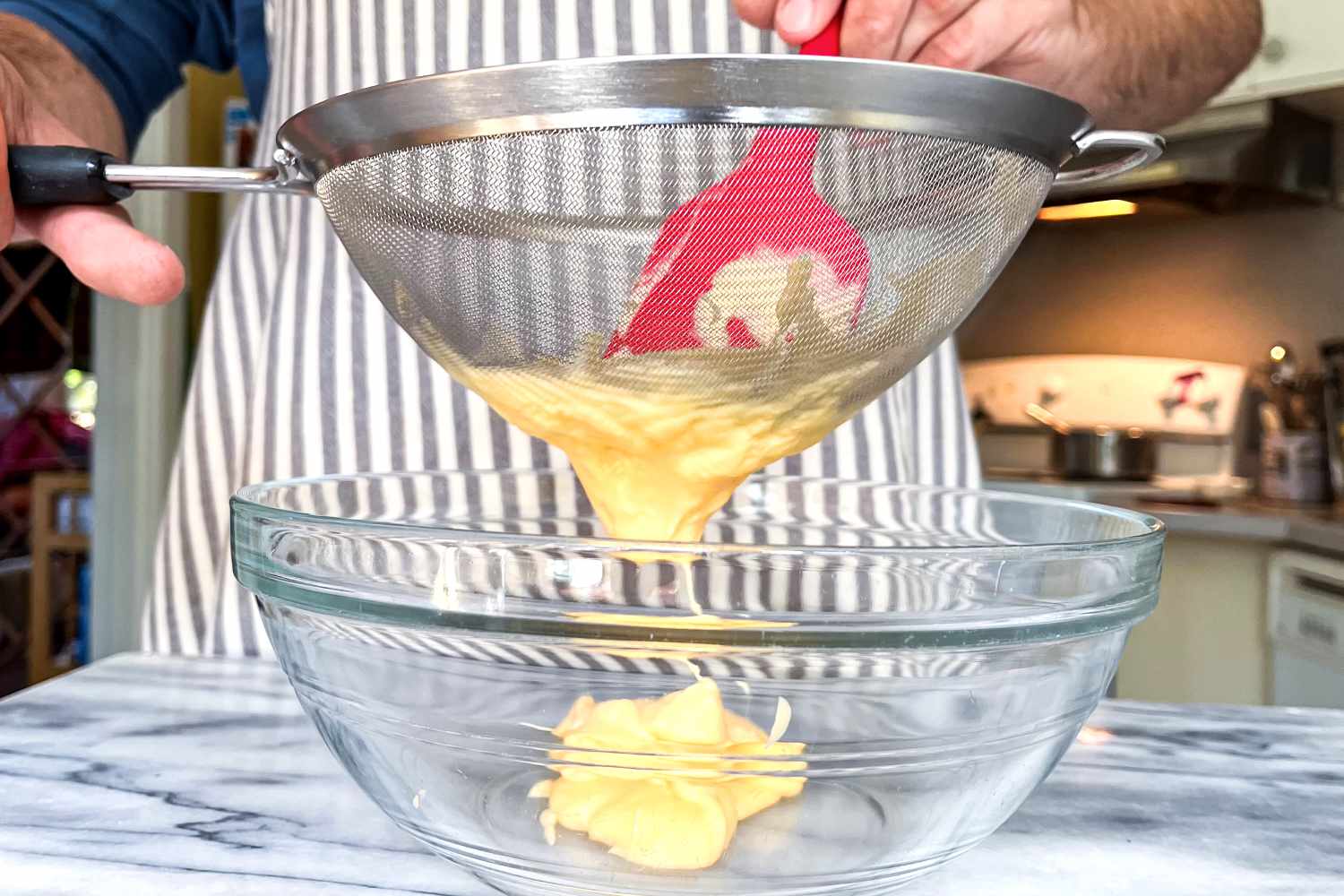 A person straining thick yellow sauce through the OXO Good Grips 8-Inch Double Rod Strainer into a glass bowl with a spatula