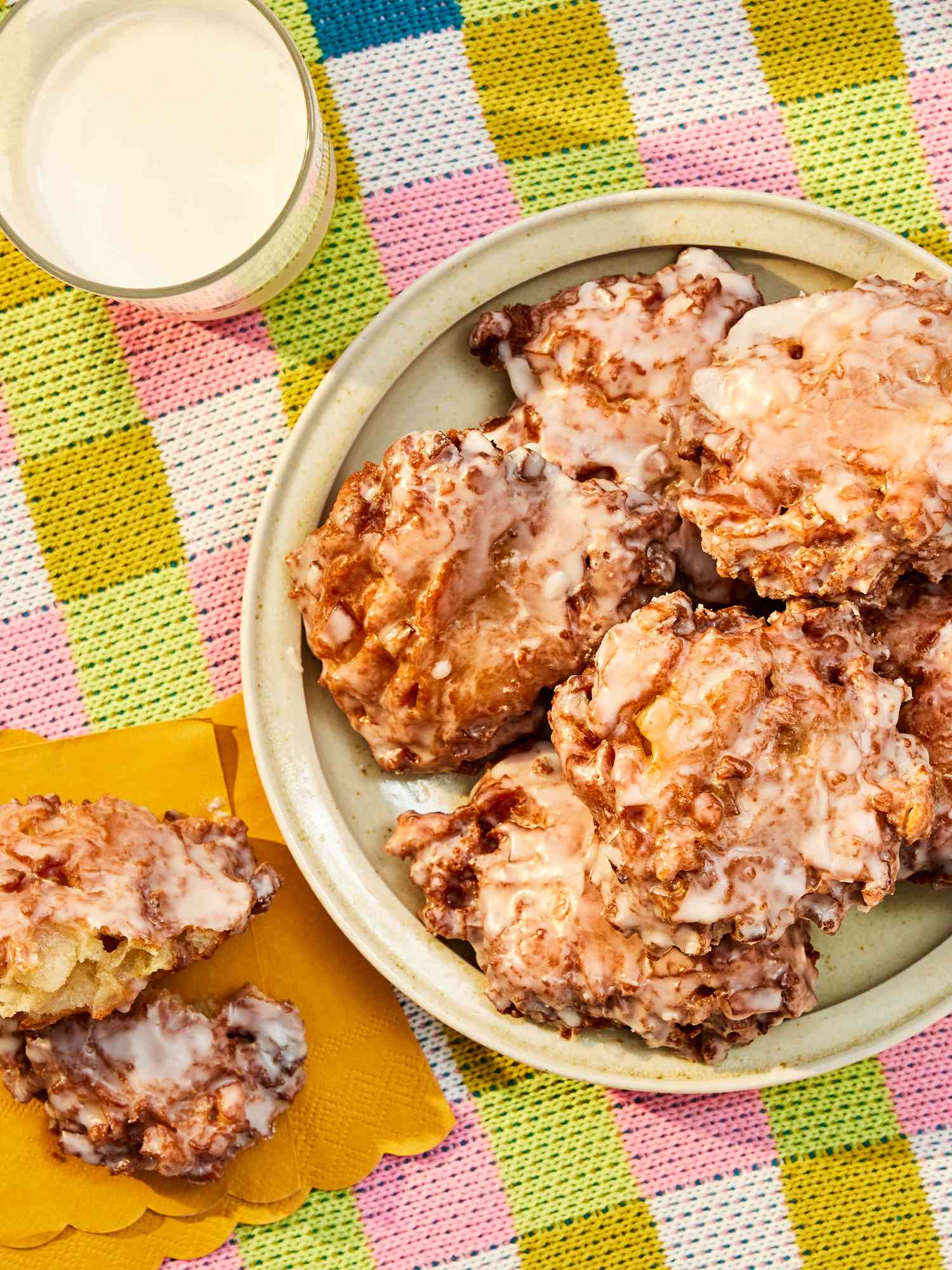 Platter of apple fritters on a round dish, with one fritter on yellow napkins, ripped open. Glass of milk, and colorful checkered tablecloth