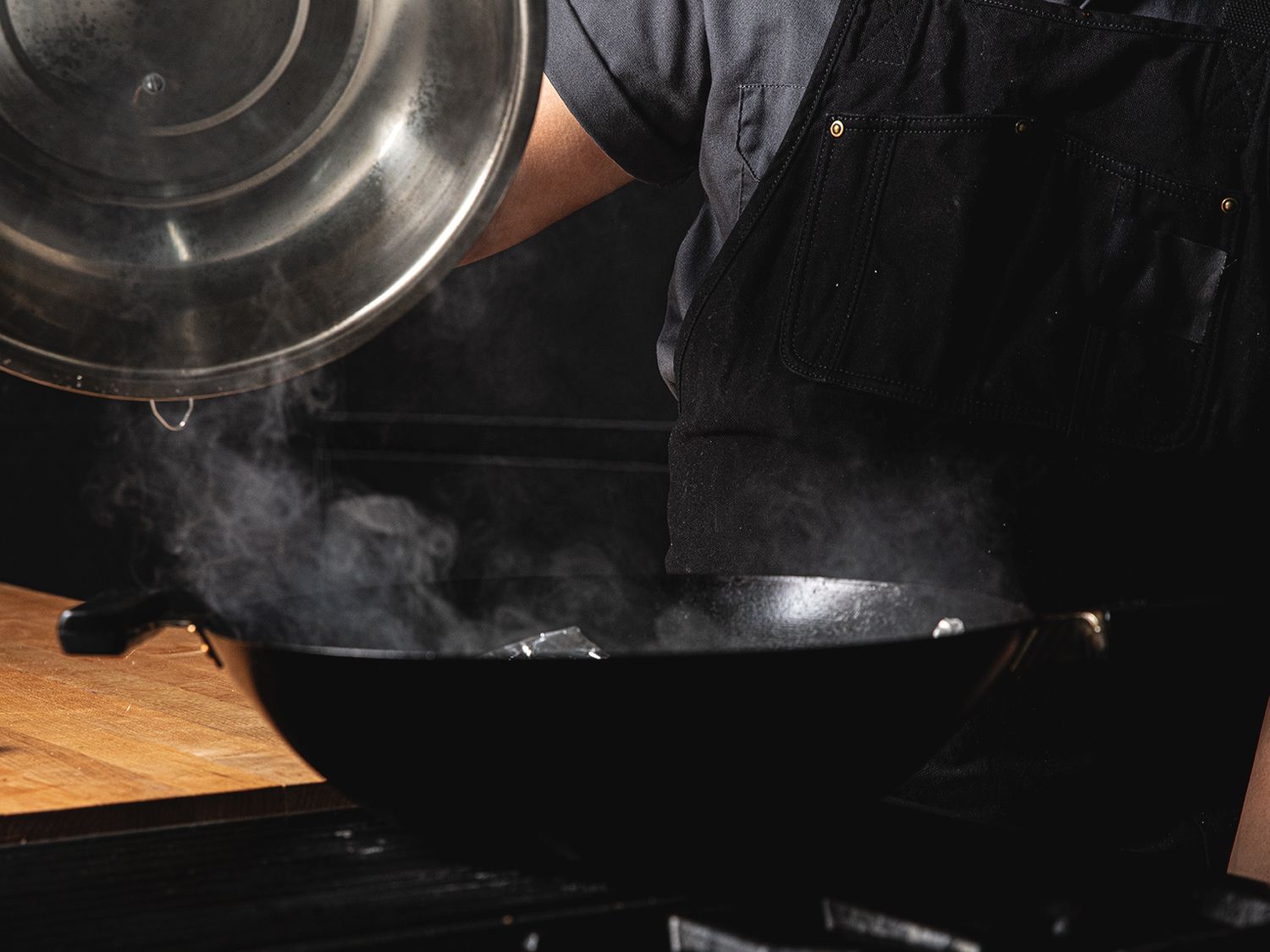 A metal lid being lifted off of a wok, with smoke escaping.