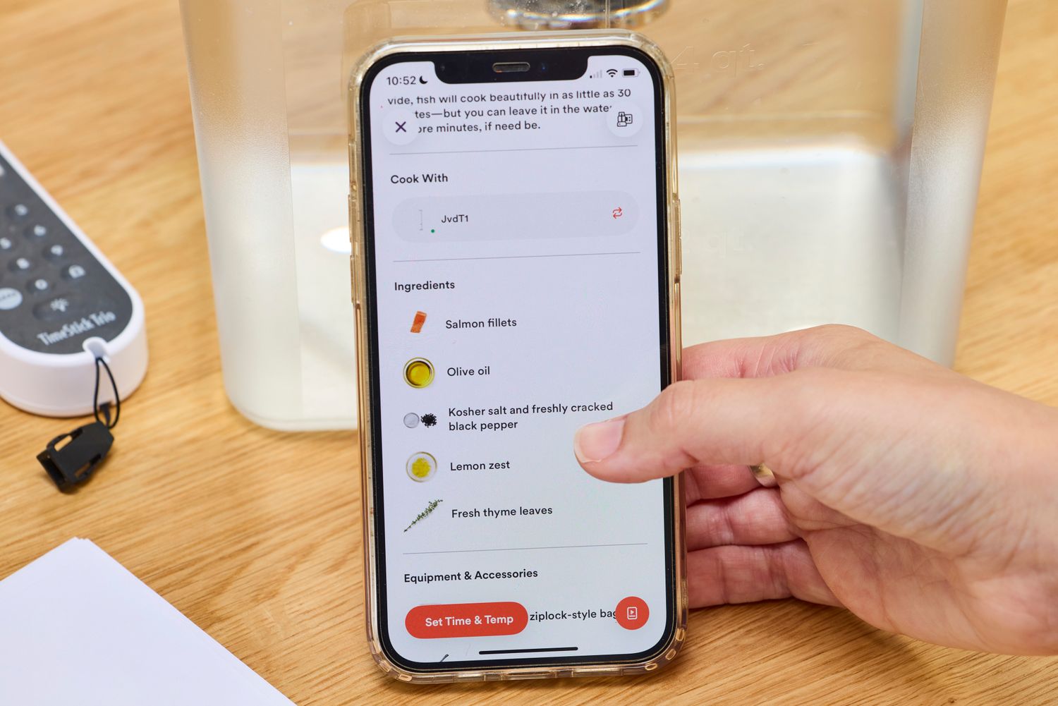 A person holding a smartphone displaying a cooking recipe on the Breville Joule Turbo Sous Vide Machine app interface, with an ingredient list including salmon and olive oil