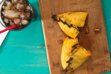 A Bolivian salteña, served on a wooden board with an ice-filled glass of cola.
