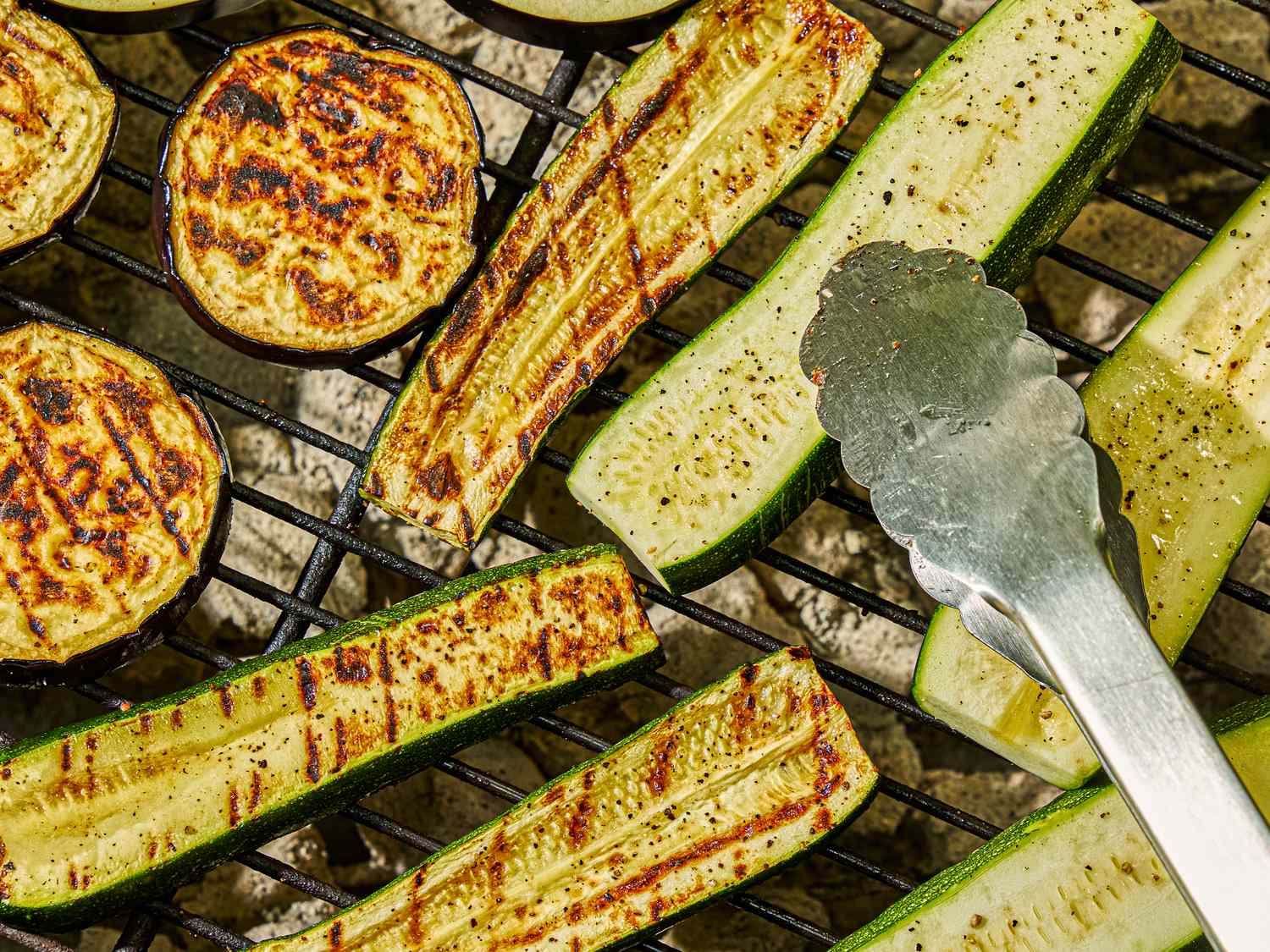 Slices of zucchini and eggplant grilling on a barbecue with metal tongs