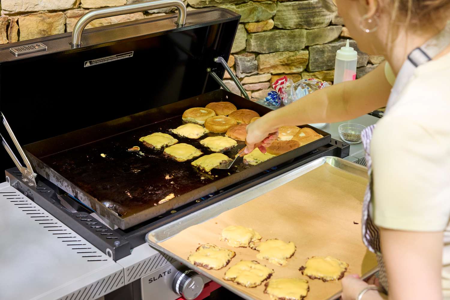 A person cooks hamburgers on the Weber Slate 28 inch Rust-Resistant 3 Burner Outdoor Griddle