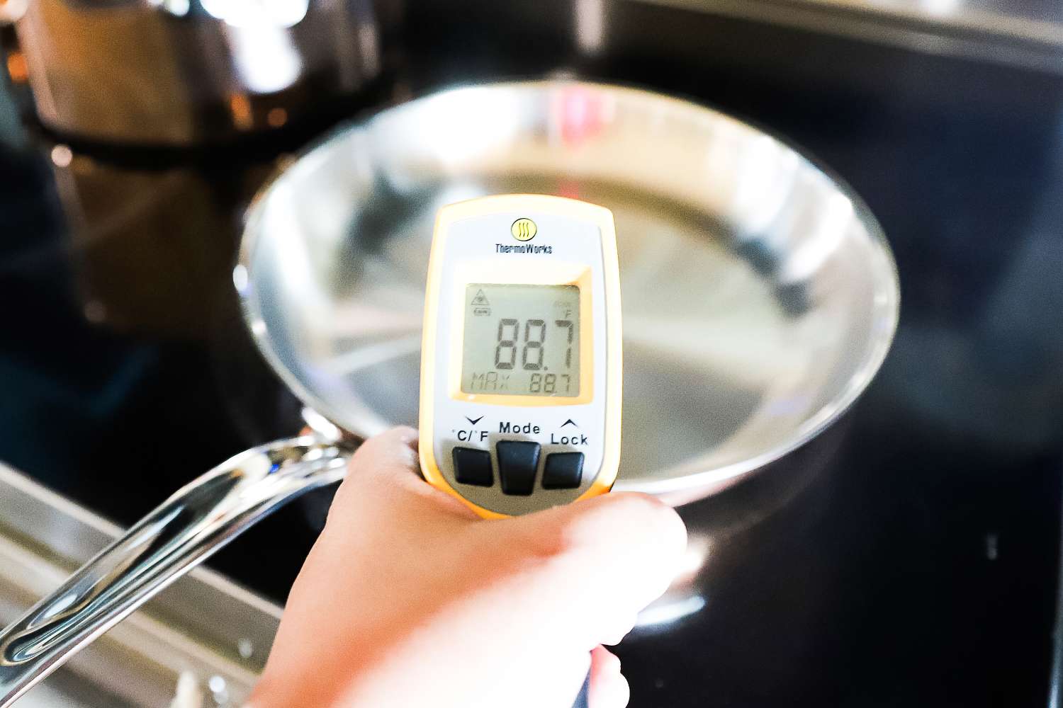 A hand holding a digital thermometer showing 887F in front of a metal frying pan