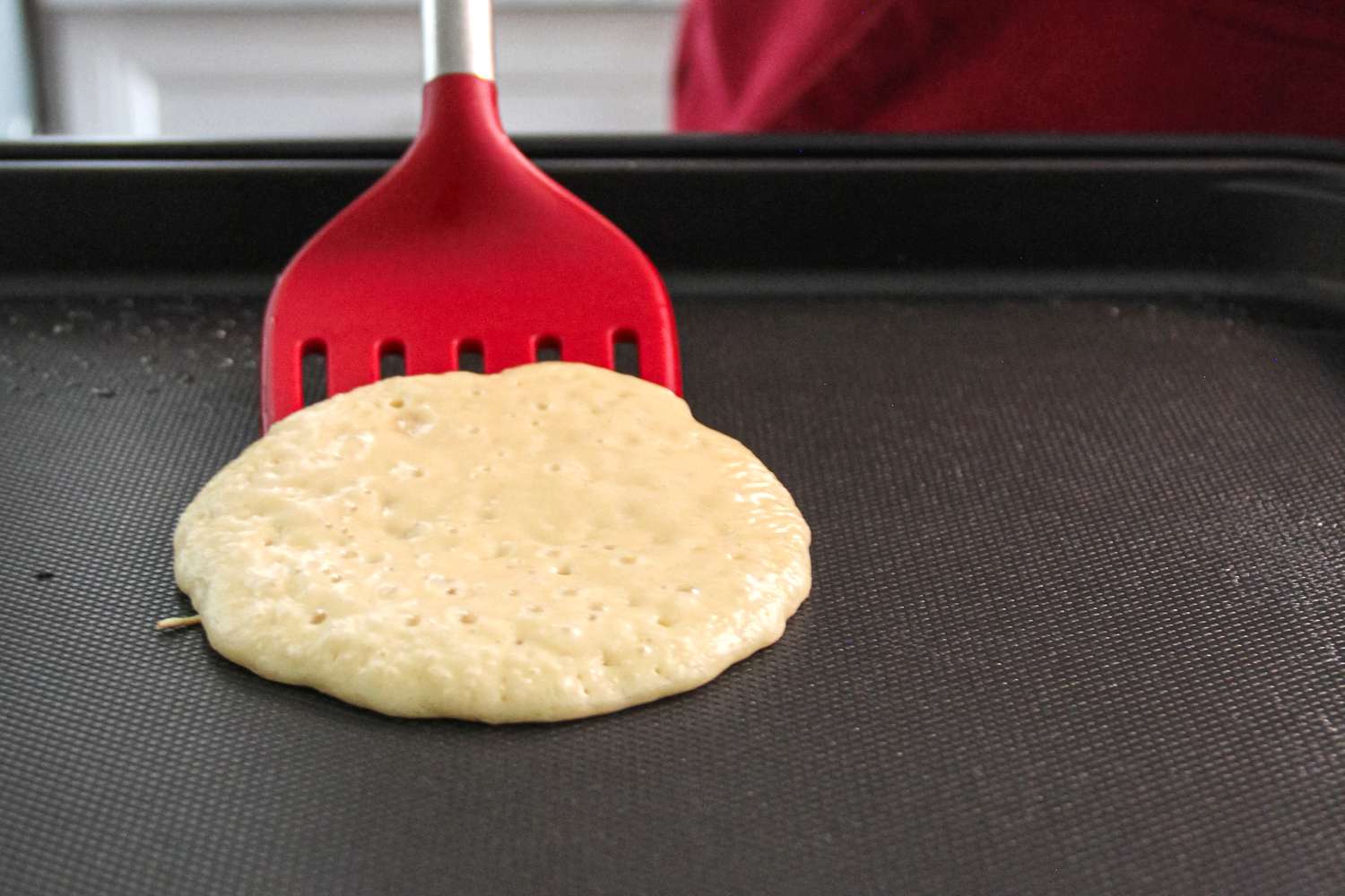 A red nonstick food turner sliding underneath an uncooked pancake on an electric griddle.