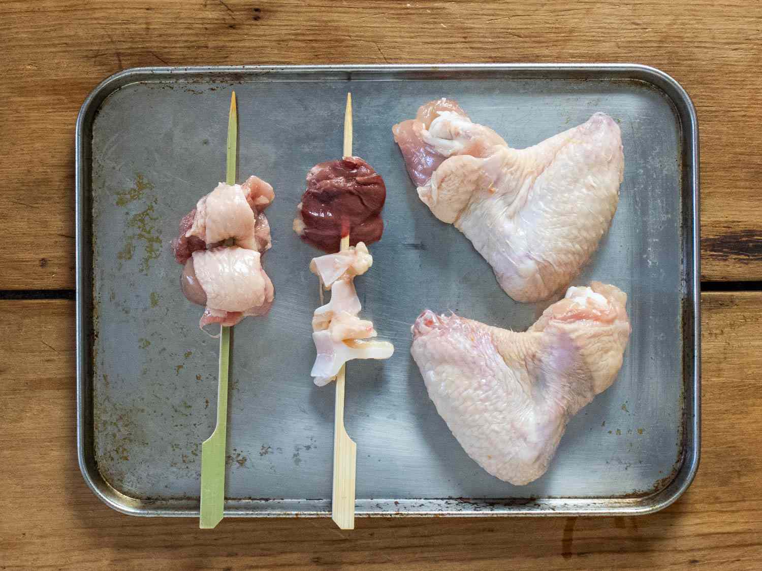 From left to right, chicken oyster on a skewer, chicken heart and cartilage on a skewer, and two chicken wings; all of them on a small sheet pan