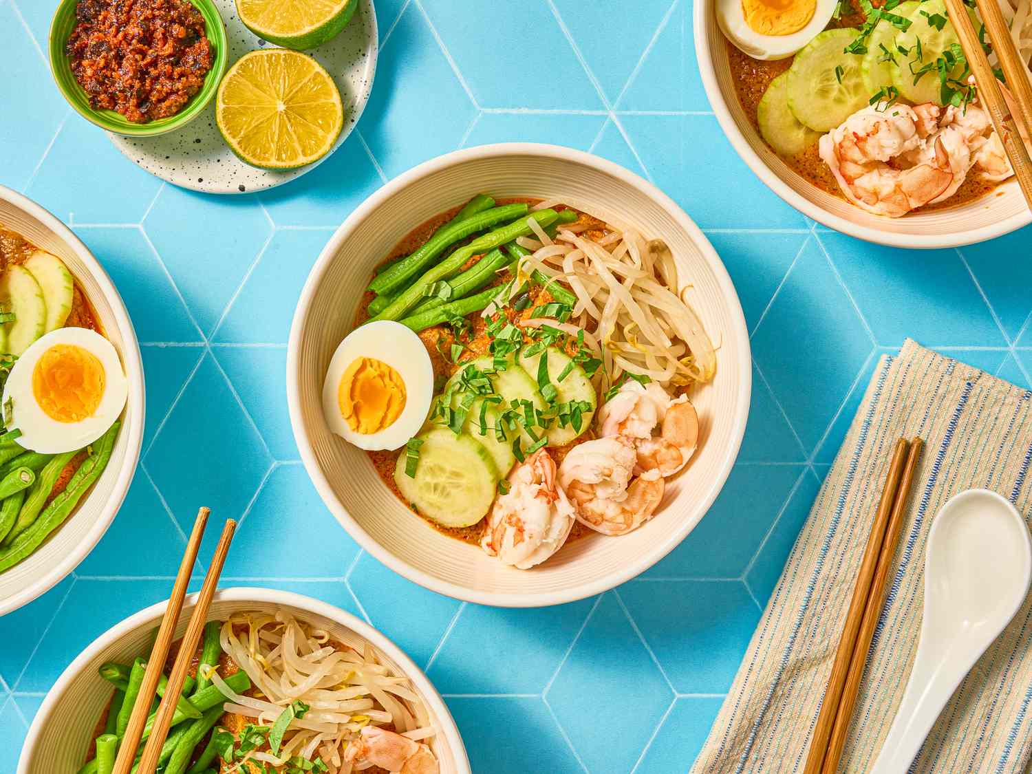 Bowls of curry laksa garnished with shrimp boiled egg cucumber and beans accompanied by lime and chili paste on a blue background
