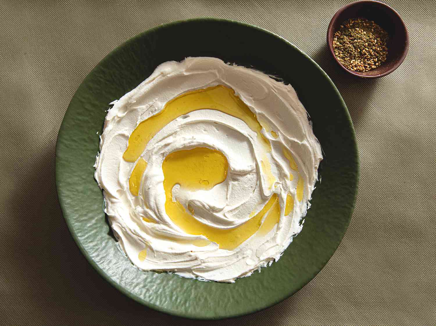 A green plate filled with labneh and oil with a small bowl of Zatar on the side