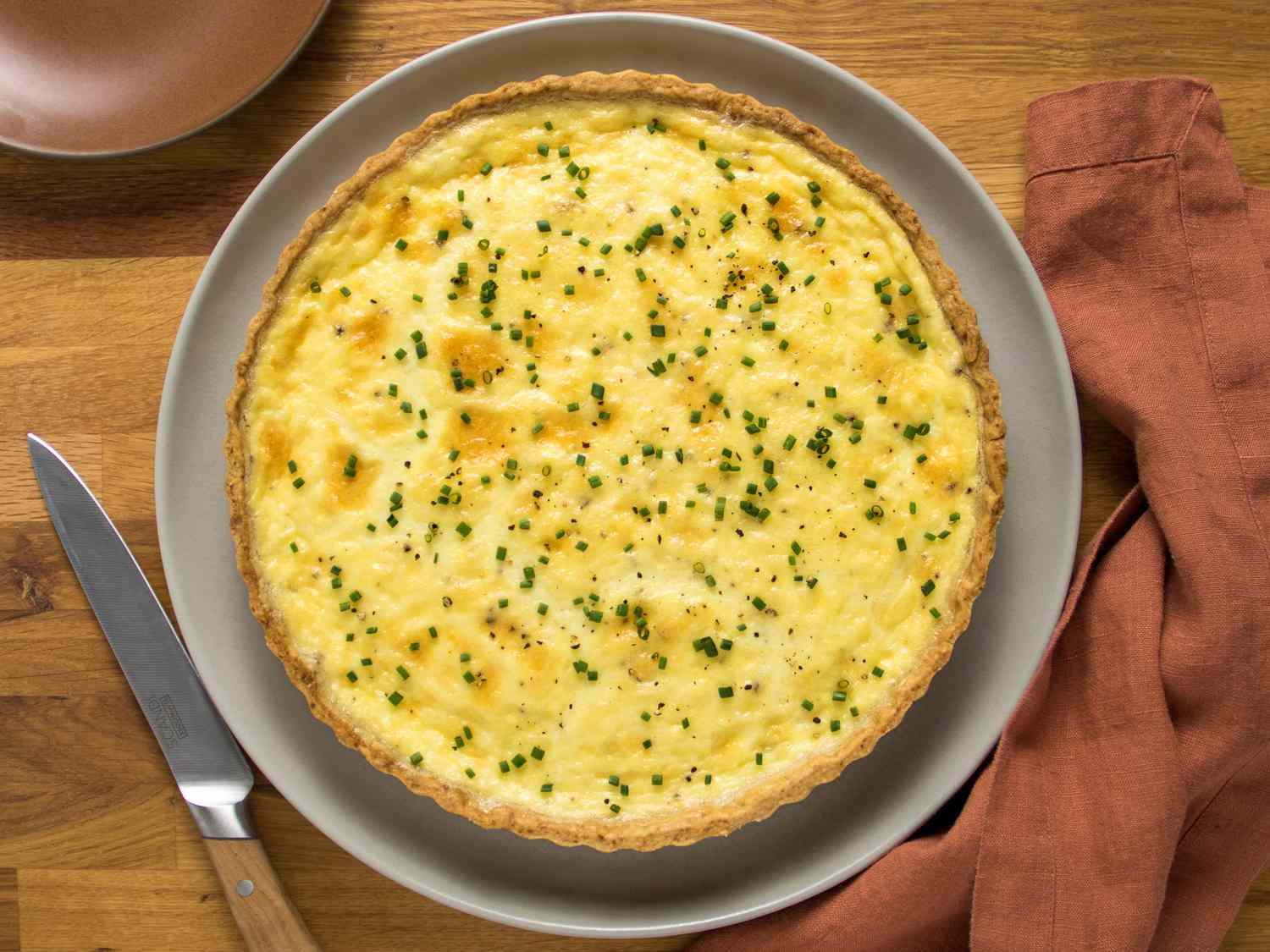 overhead shot of whole cheese quiche on a wooden tabletop, with a brick colored napkin and plate on each side. 