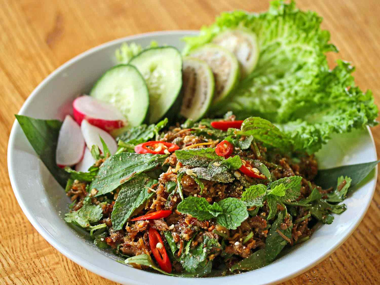 A bowl of laab ped, a salad of ground duck meat, chilies, and herbs, topped with whole mint leaves, with lettuce, cucumber, and radish on the side.